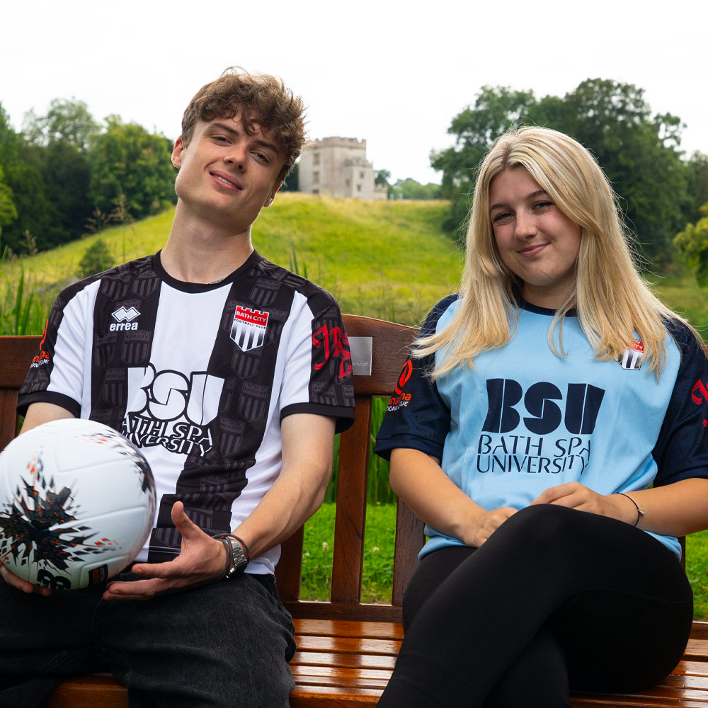 Two young people sitting on a bench in Bath City football tops with BSU logos across the front
