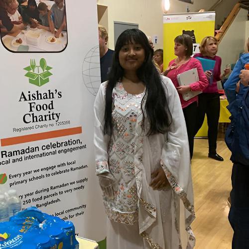 Student Aishah Ahmed stands next to a banner that says Aisha's Food Charity