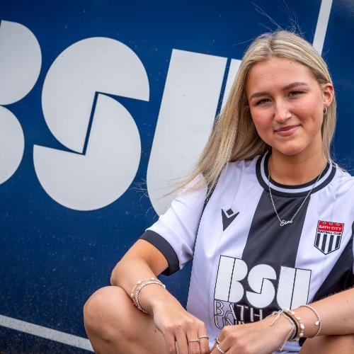 BSU Student Esme wearing a black and white Bath City shirt in front of a big BSU logo