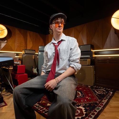A student wearing a shirt and tie in a music room, with large amp speakers behind him