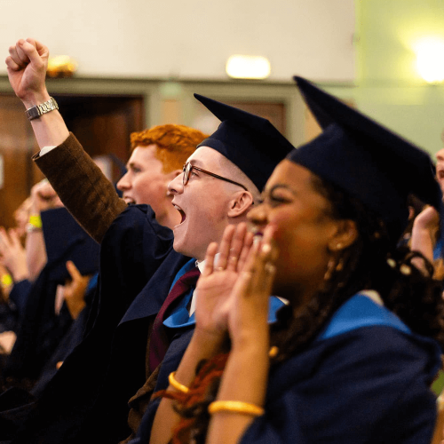 Two rows of students at their final year graduation looking at the stage and cheering.